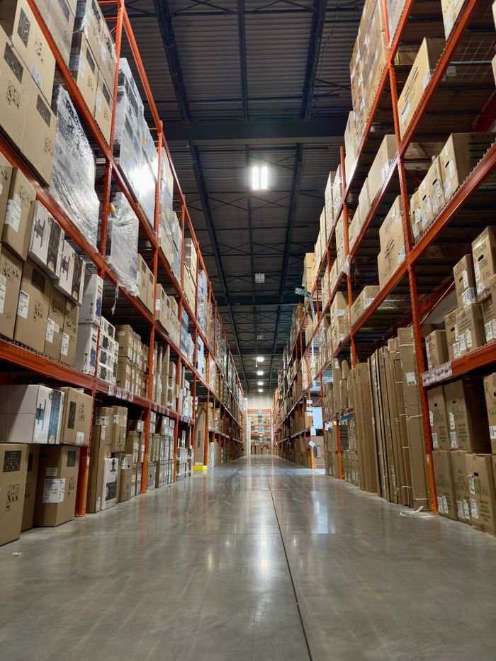 About Long aisle in a busy warehouse stacked with boxes and products on shelves.