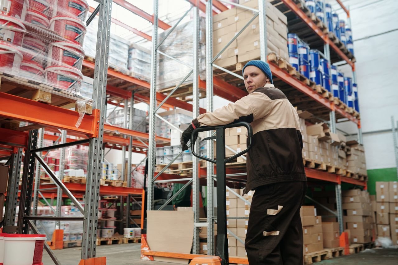 our-journey-02 Warehouse worker operating a pallet jack among stacked boxes and metal shelves in an industrial setting.